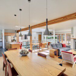 Open-concept living space with a long wooden dining table, pendant lights, and a kitchen area with stainless appliances alongside a gray sofa and fireplace.