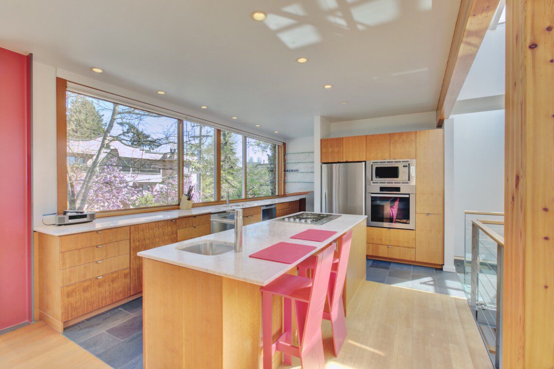 Bright modern kitchen with wooden cabinets, a marble island, pink chairs, and large windows overlooking trees outside.