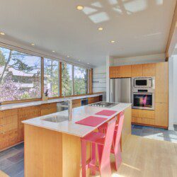Bright modern kitchen with wooden cabinets, a marble island, pink chairs, and large windows overlooking trees outside.