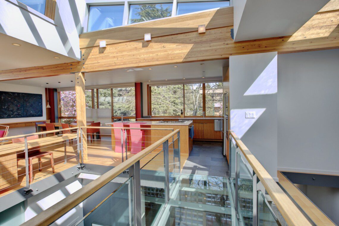 Open-concept kitchen and dining area in a modern home with wood beams, glass railing, and pink chairs near a large windowed wall.