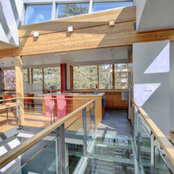 Open-concept kitchen and dining area in a modern home with wood beams, glass railing, and pink chairs near a large windowed wall.