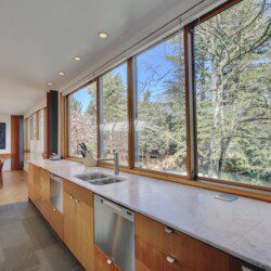 Bright kitchen with marble countertop, wooden cabinets, stainless sink and dishwasher, and large windows overlooking trees outside.