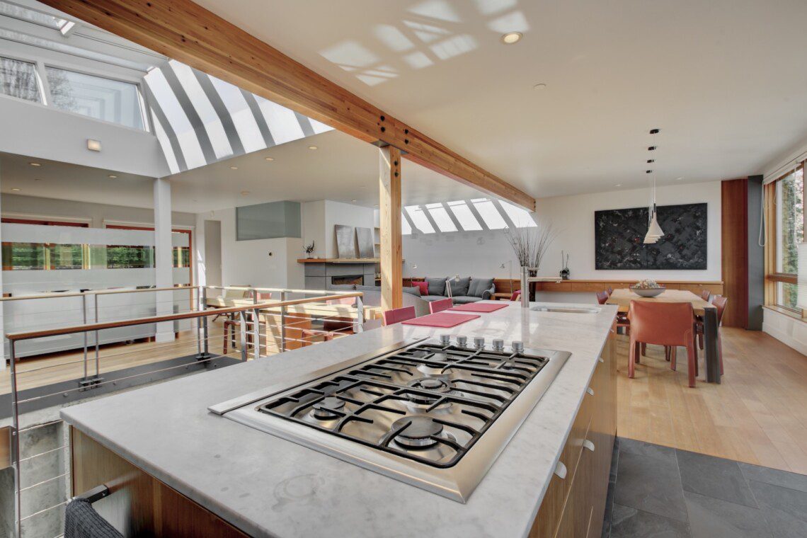 Open-plan kitchen with marble island and stainless gas range, wooden beams, and bright dining area with red chairs under skylights.