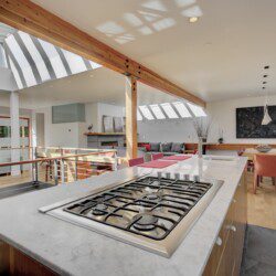 Open-plan kitchen with marble island and stainless gas range, wooden beams, and bright dining area with red chairs under skylights.