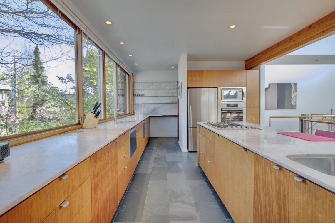 Bright, sunlit kitchen with long marble counters, oak cabinets, and stainless steel fridge and oven, seen through large windows.