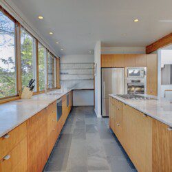 Bright, sunlit kitchen with long marble counters, oak cabinets, and stainless steel fridge and oven, seen through large windows.