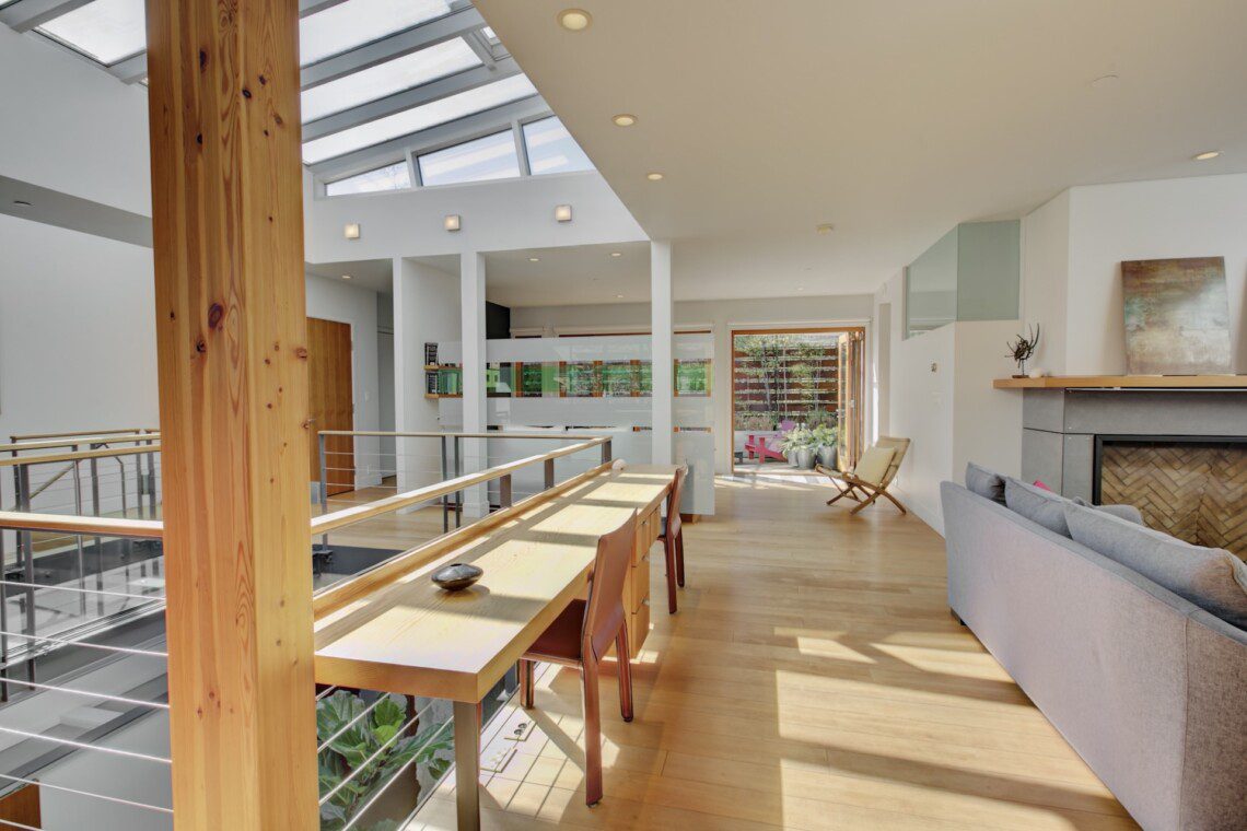 Bright open-plan living area with skylights, a tall wooden pillar, and a long wooden dining table by a glass railing.