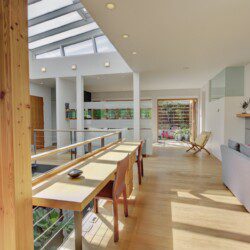 Bright open-plan living area with skylights, a tall wooden pillar, and a long wooden dining table by a glass railing.
