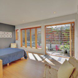 Bright bedroom with a blue bedspread, wooden floor, and wicker chair overlooking a patio through large wooden-framed windows and glass doors.