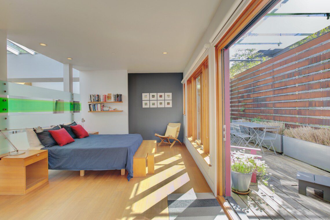Bright modern bedroom with a blue bed, red and navy pillows, and wooden furniture, opening to a sunny patio through large glass doors.