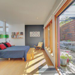 Bright modern bedroom with a blue bed, red and navy pillows, and wooden furniture, opening to a sunny patio through large glass doors.