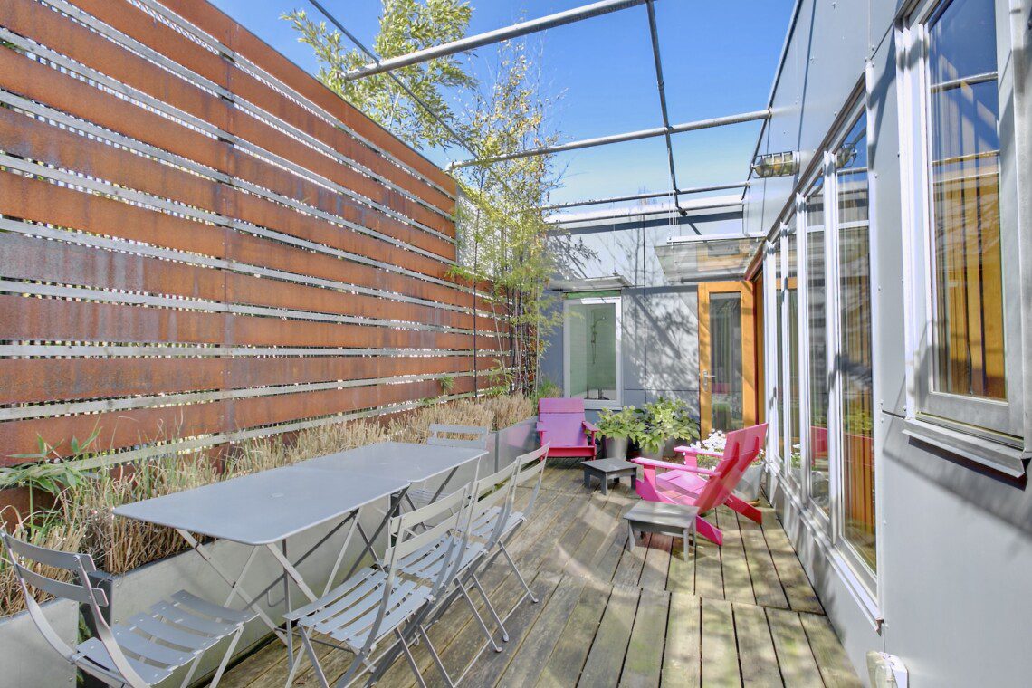 Rooftop patio with a long grey table and metal folding chairs beside a rust-colored privacy wall; pink chairs, plants, and a glass-walled building.