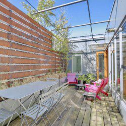 Rooftop patio with a long grey table and metal folding chairs beside a rust-colored privacy wall; pink chairs, plants, and a glass-walled building.