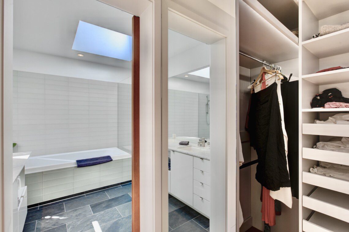 Bright bathroom with a built-in bathtub, white tiled walls, and a blue towel draped over the tub, seen through a doorway into a mirrored vanity area.