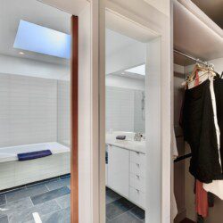 Bright bathroom with a built-in bathtub, white tiled walls, and a blue towel draped over the tub, seen through a doorway into a mirrored vanity area.