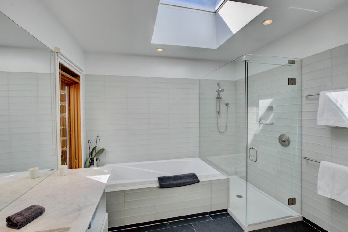 Bright modern bathroom with a white tiled bath and glass-enclosed shower under a skylight.