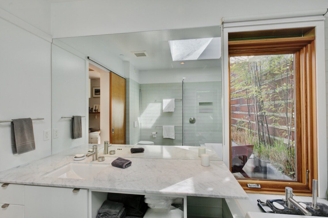 Bright, modern bathroom with a marble double-sink vanity, large wall mirror, gray towels, and a glass shower enclosure adjacent to a wooden-framed window. Outside green plants are visible through the window, adding natural light to the space.