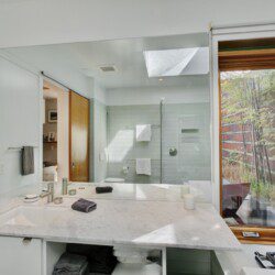 Bright, modern bathroom with a marble double-sink vanity, large wall mirror, gray towels, and a glass shower enclosure adjacent to a wooden-framed window. Outside green plants are visible through the window, adding natural light to the space.