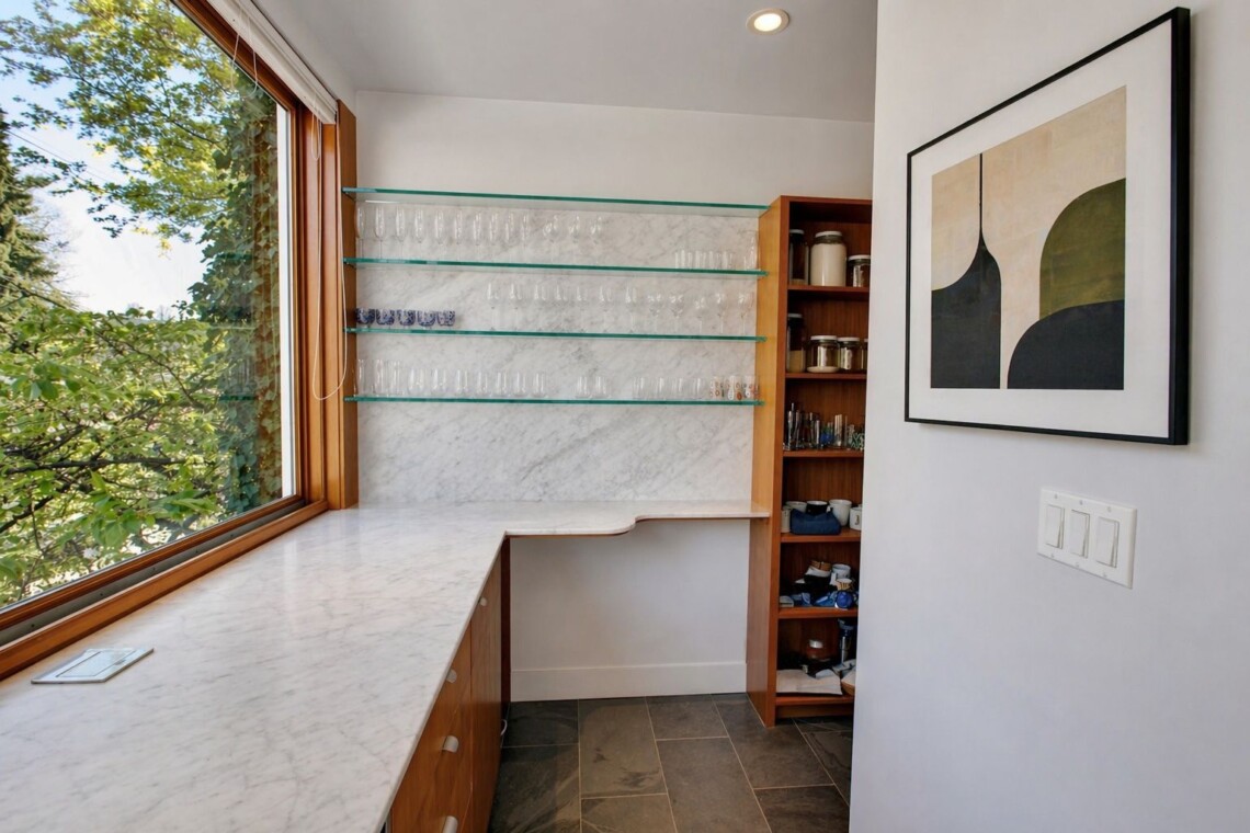 Home bar nook with marble countertop, glass shelves holding stemware, and a wooden cabinet near a large window.