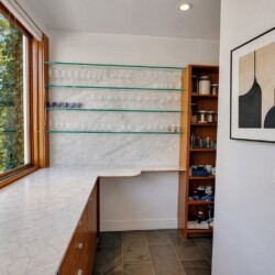 Home bar nook with marble countertop, glass shelves holding stemware, and a wooden cabinet near a large window.
