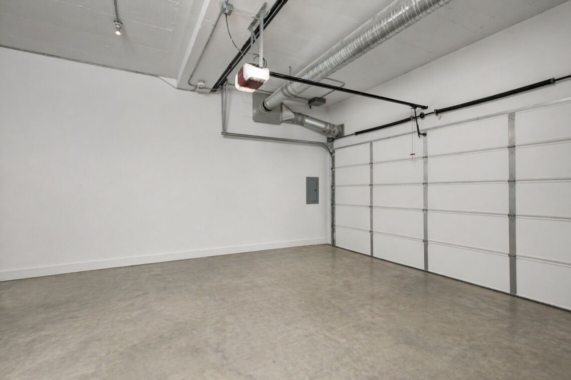 Empty residential garage with a large white sectional door and exposed ceiling ducts and wiring, concrete floor.