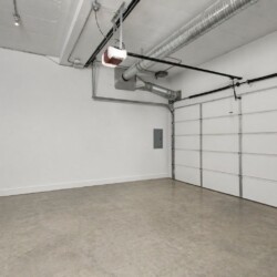 Empty residential garage with a large white sectional door and exposed ceiling ducts and wiring, concrete floor.