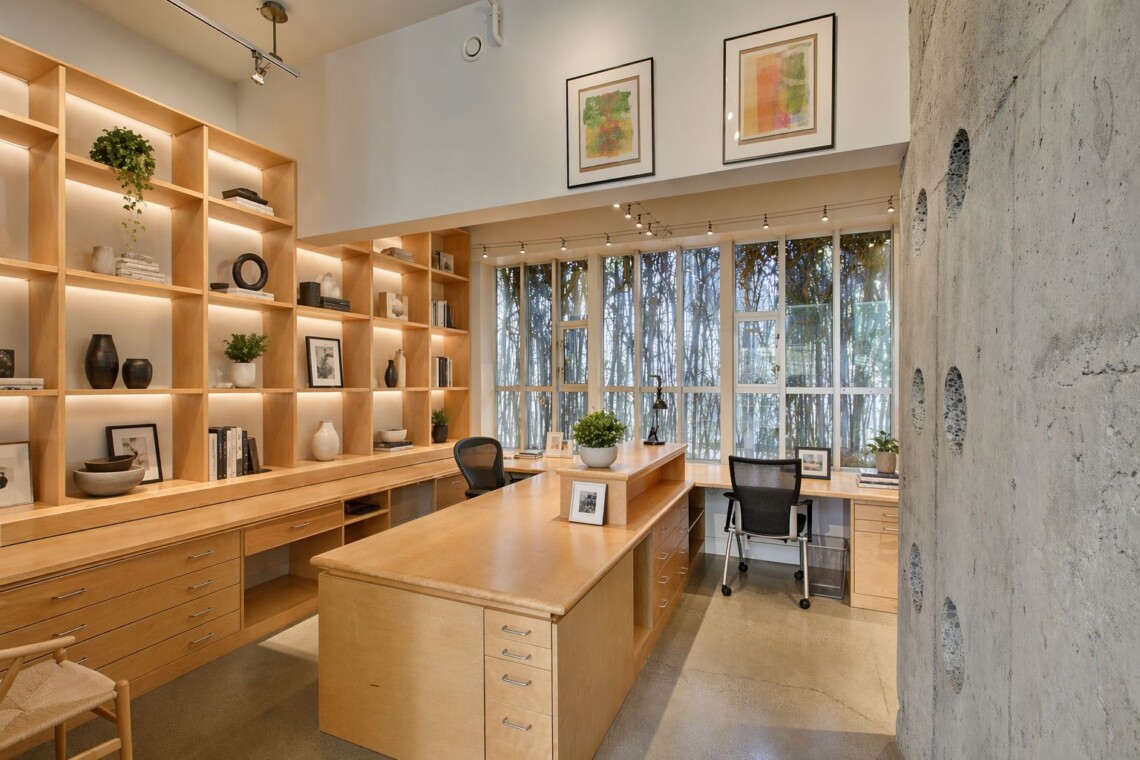 Modern office with wooden built-in shelves, warm lighting, and a central wooden island workstation surrounded by chairs.