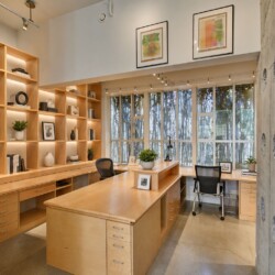 Modern office with wooden built-in shelves, warm lighting, and a central wooden island workstation surrounded by chairs.