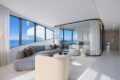 Bright, modern living room with curved floor-to-ceiling windows showing a blue ocean view and white architectural curves outside.