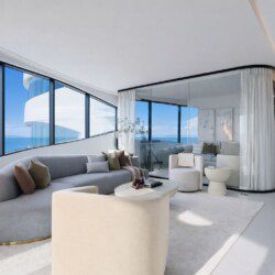 Bright, modern living room with curved floor-to-ceiling windows showing a blue ocean view and white architectural curves outside.