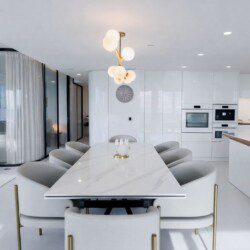 Luxurious open-plan dining and kitchen area with a marble table, curved grey chairs, glossy white cabinets, and a brass chandelier, overlooking a sea view through glass walls.