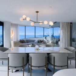 Open-concept dining and living area with a marble table, cream chairs, and a panoramic ocean view through large glass windows.
