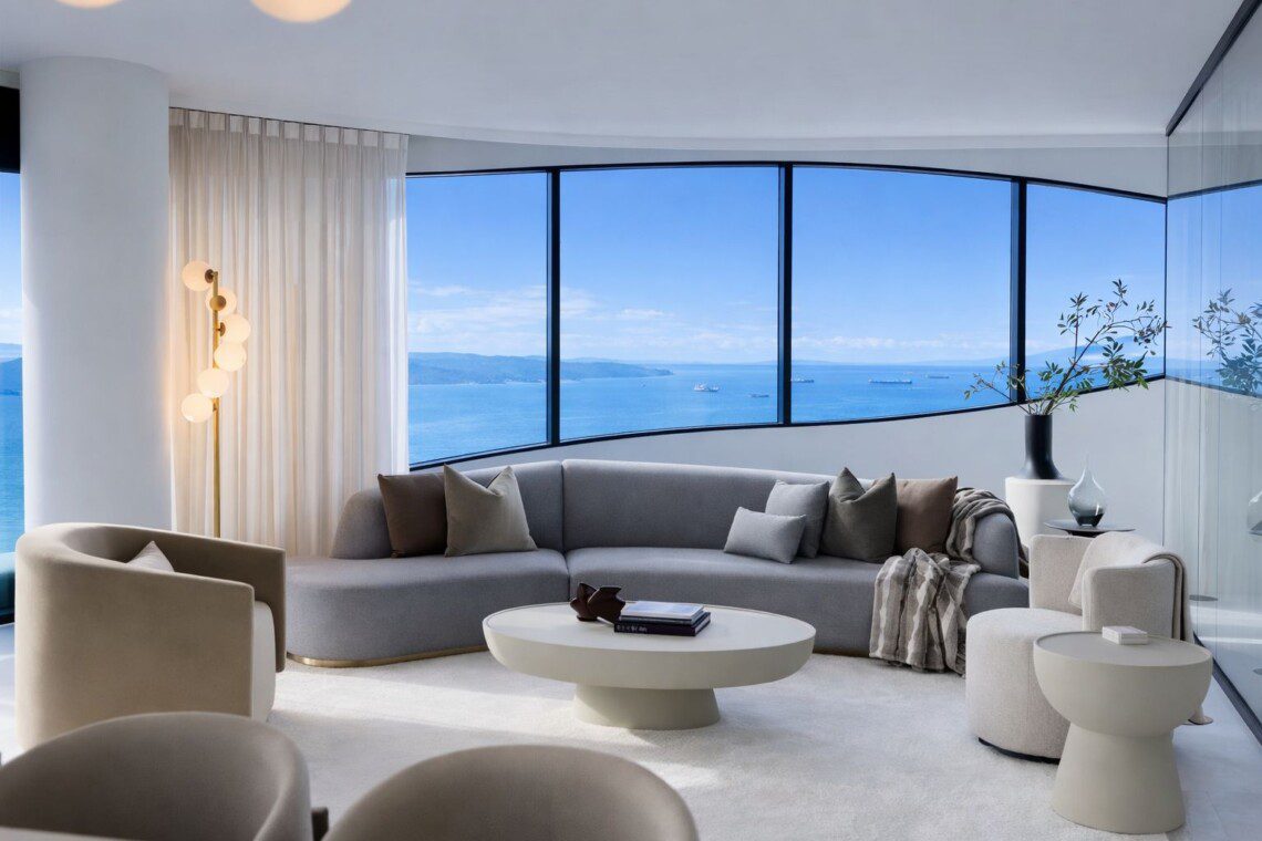 Bright modern living room with a curved window wall showing the blue ocean and ships; grey sectional sofa, beige chairs, and a white circular coffee table.