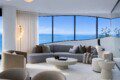 Bright modern living room with a curved window wall showing the blue ocean and ships; grey sectional sofa, beige chairs, and a white circular coffee table.
