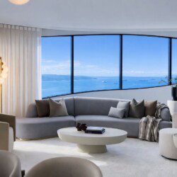 Bright modern living room with a curved window wall showing the blue ocean and ships; grey sectional sofa, beige chairs, and a white circular coffee table.