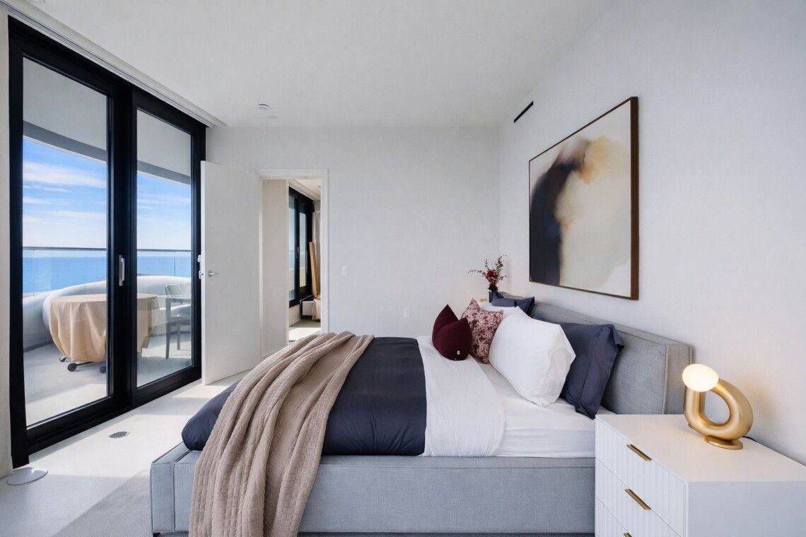 Bright modern bedroom with floor-to-ceiling glass doors opening to a balcony with an ocean view; bed with white and navy bedding and a beige throw.