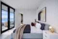 Bright modern bedroom with floor-to-ceiling glass doors opening to a balcony with an ocean view; bed with white and navy bedding and a beige throw.