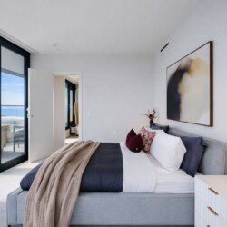 Bright modern bedroom with floor-to-ceiling glass doors opening to a balcony with an ocean view; bed with white and navy bedding and a beige throw.