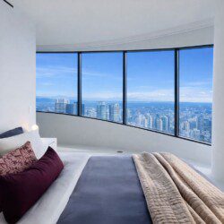 Bed in a minimalist hotel room with burgundy pillows and a beige quilt, facing curved floor-to-ceiling windows overlooking a city skyline.
