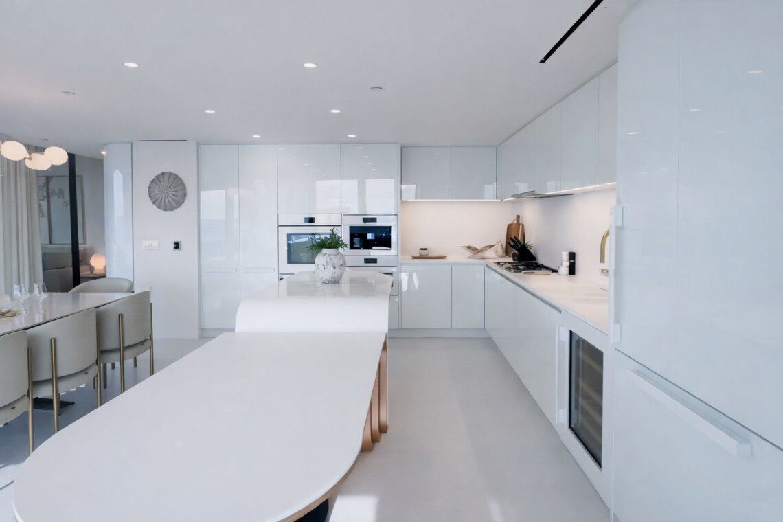 Bright white kitchen with glossy cabinets, an oval island, and built-in double ovens in the background.