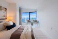 Hotel-style bedroom with a large window seating area overlooking a city skyline; bed in foreground with neutral bedding and pillows.