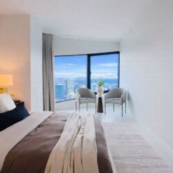 Hotel-style bedroom with a large window seating area overlooking a city skyline; bed in foreground with neutral bedding and pillows.