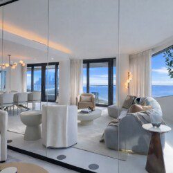Bright open-concept living room with floor-to-ceiling windows and an ocean view, neutral sofas, round coffee tables, and soft beige decor.