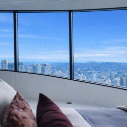 Modern bedroom with a curved wall of floor-to-ceiling windows overlooking a clear blue city skyline.