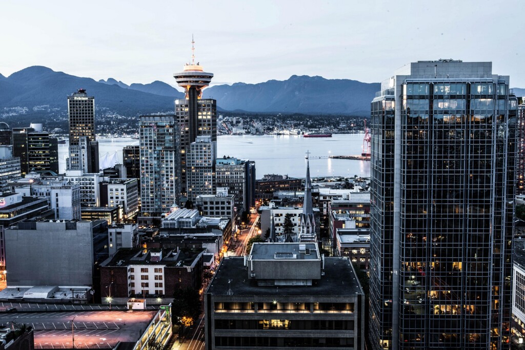 City skyline along a waterfront with tall glass buildings and distant blue mountains in the background at dusk.
