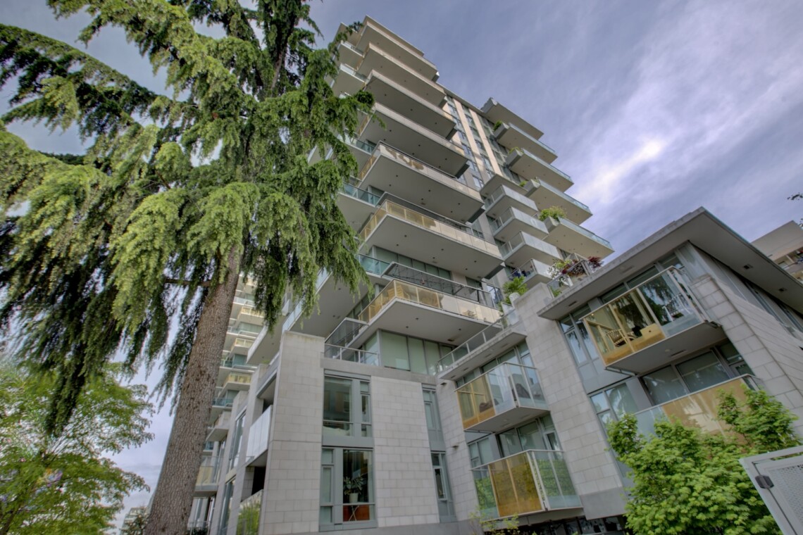 Tall modern apartment building with numerous balconies beside a large green tree under a blue sky, angled upward view