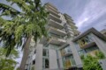 Tall modern apartment building with numerous balconies beside a large green tree under a blue sky, angled upward view