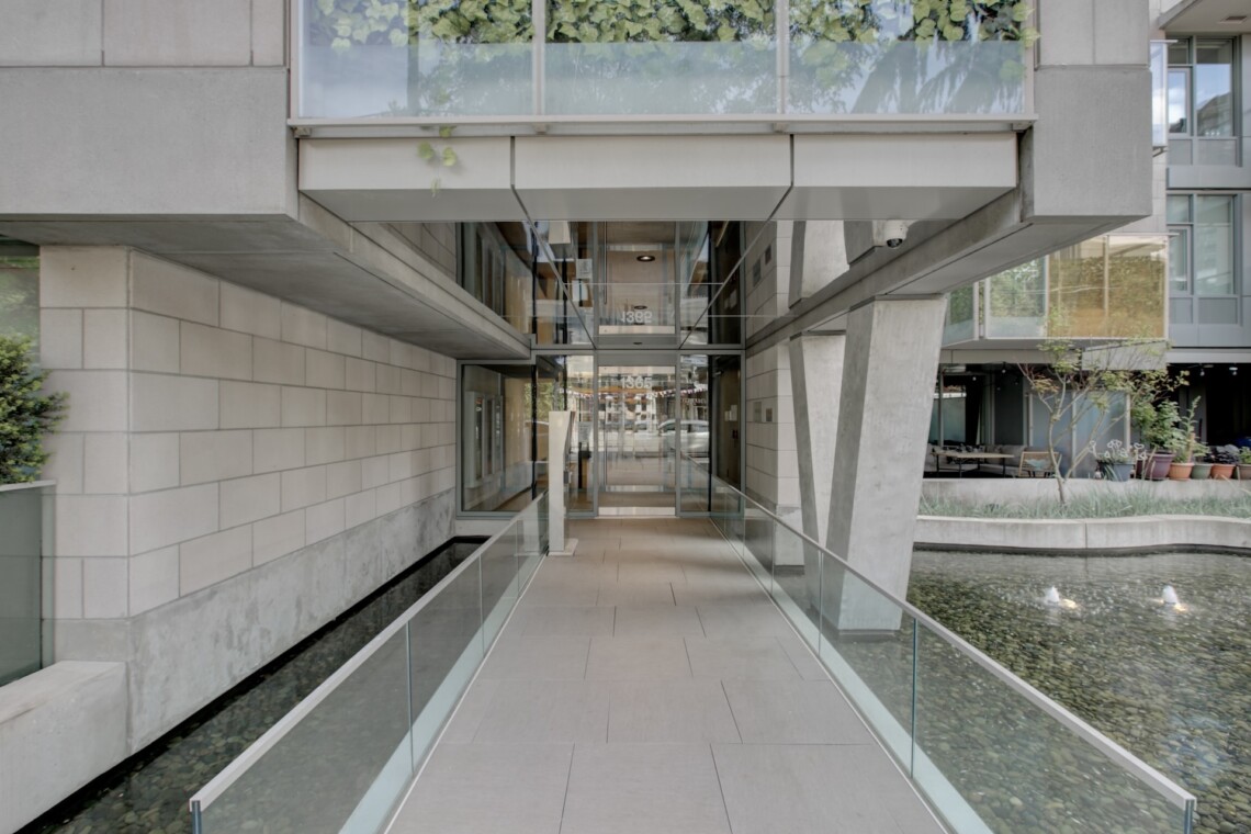 Modern concrete building entrance with glass railings and a water-filled canal on both sides, leading to glass doors at the center.