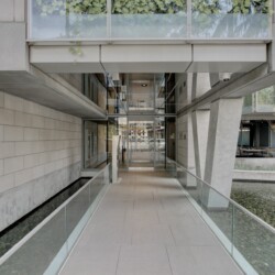 Modern concrete building entrance with glass railings and a water-filled canal on both sides, leading to glass doors at the center.