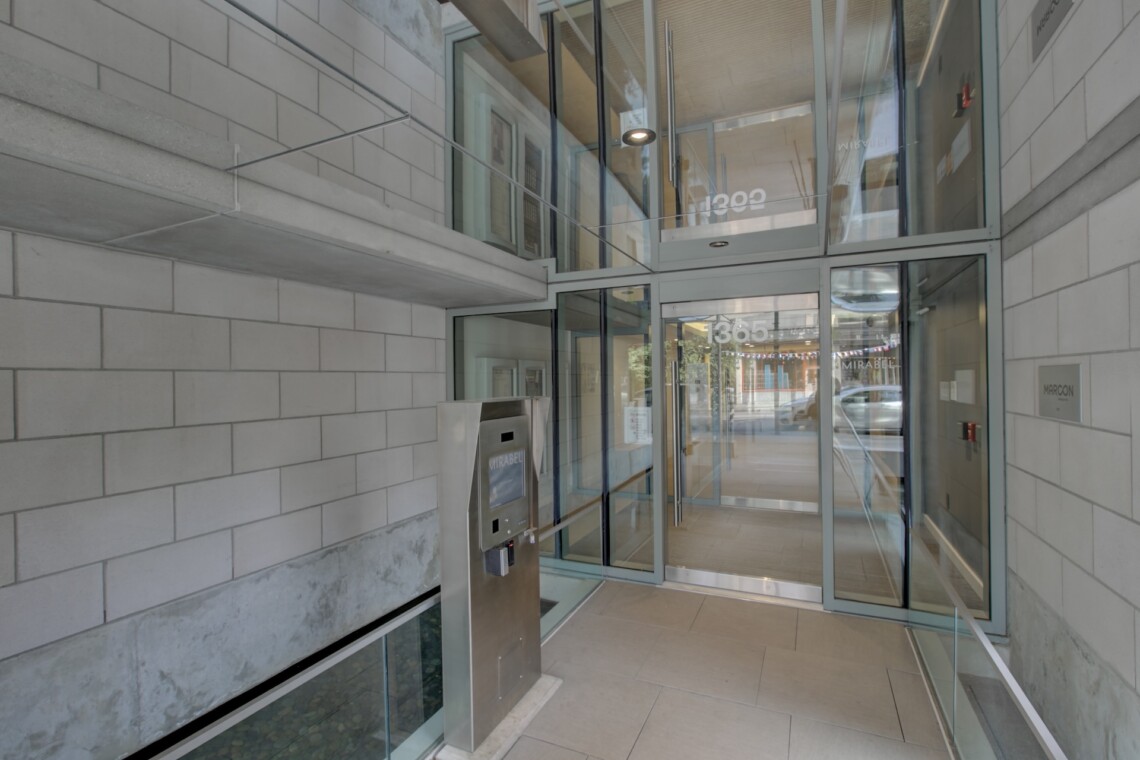Modern building entrance with large glass doors, gray brick walls, and a stainless steel security kiosk near the door.